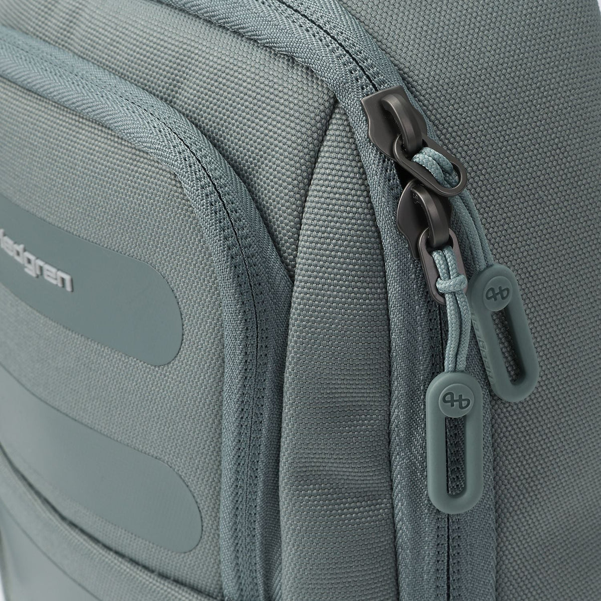 Muted grey sling bag by Hedgren with dark metal zippers, light teal cord loops, and rubber 'hb' logo pulls.