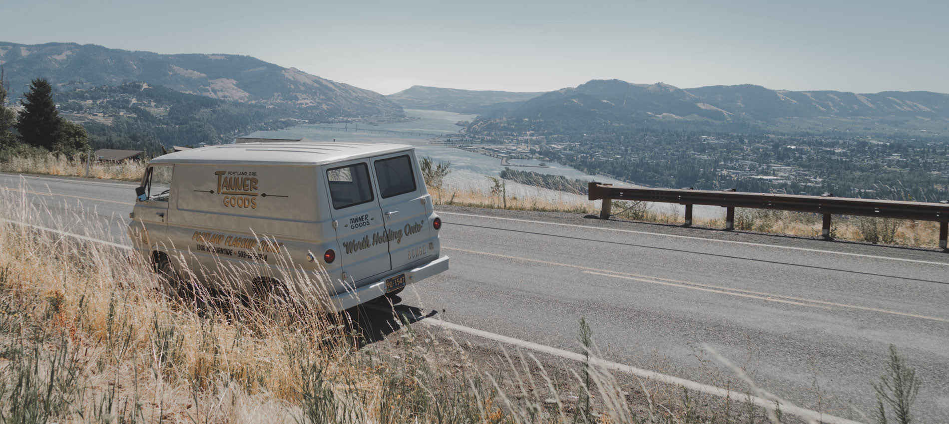 A white conversion van with Tanner Goods decals parked on the side of the road overlooking the Columbia River Gorge.