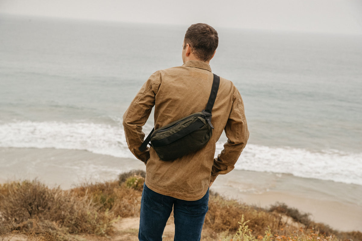 Man wearing olive green waist bag by ocean, rugged outdoor style, earthy tones, adventure-ready.