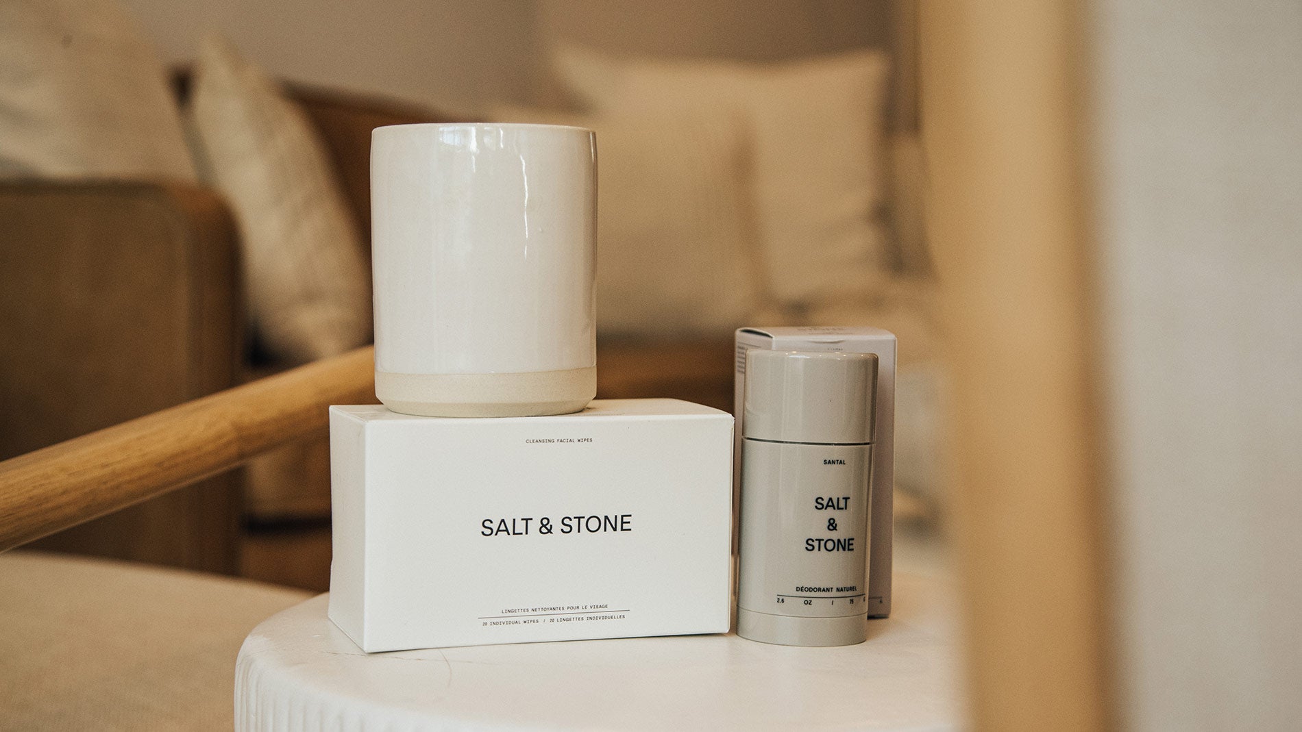 A Salt & Stone deodorant stick with a candle sitting on top of a clean, white end table.