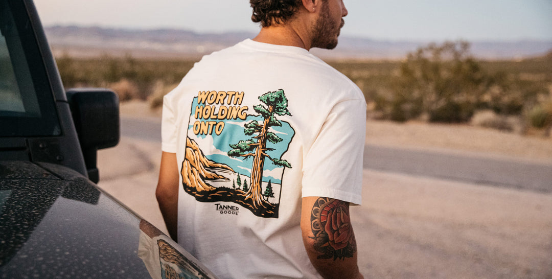 Man wearing a Tanner Goods "Worth Holding Onto" graphic t-shirt leaning up against a Jeep on the side of a desert road.
