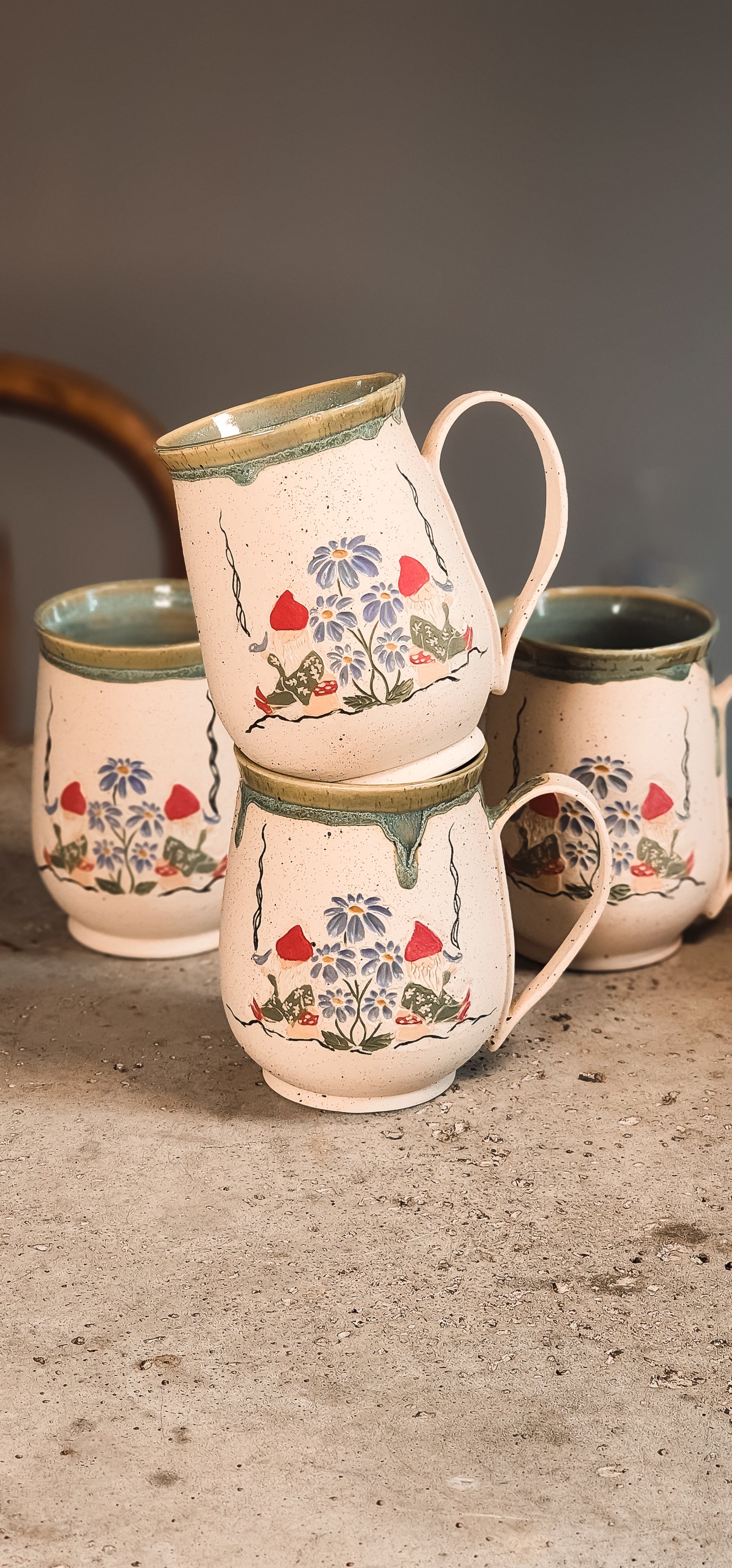 Light cream ceramic gnome mugs with hand-painted gnomes and blue flowers, stacked on a textured surface.