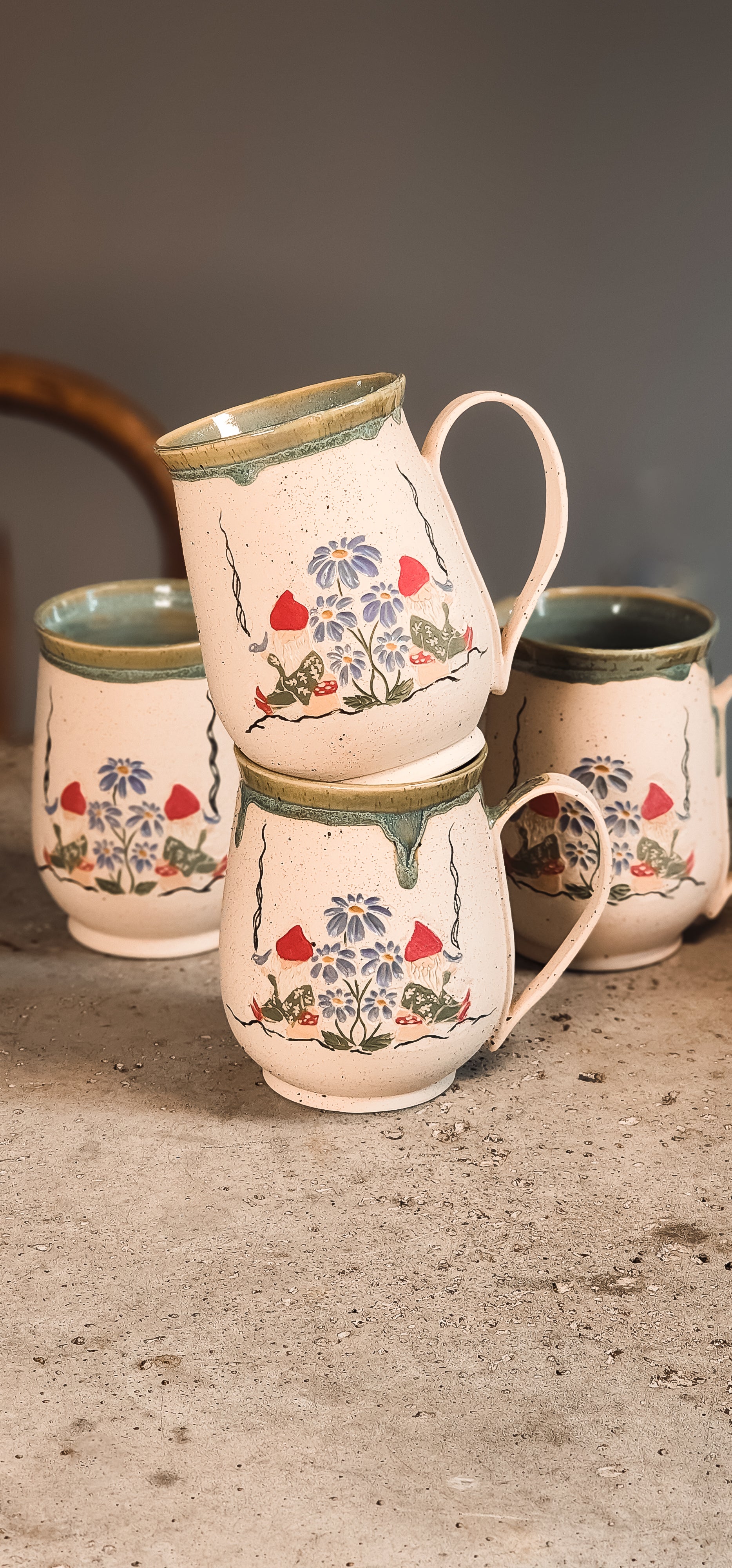 Light cream ceramic gnome mugs with hand-painted gnomes and blue flowers, stacked on a textured surface.