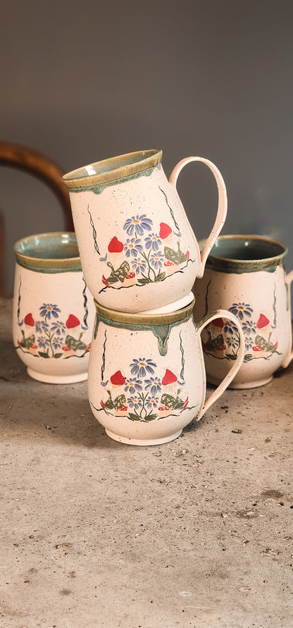 Light cream ceramic gnome mugs with hand-painted gnomes and blue flowers, stacked on a textured surface.