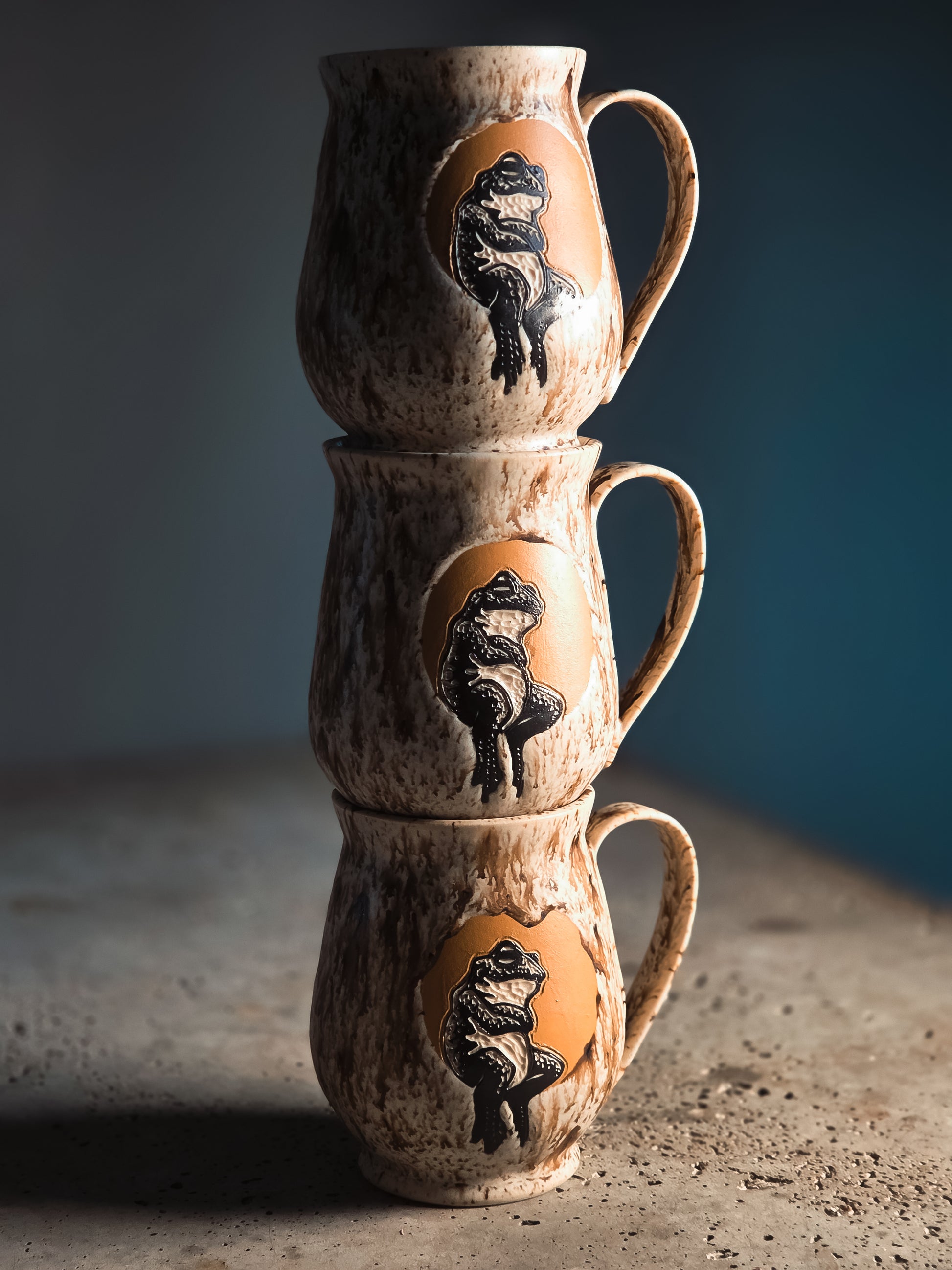 Three stacked brown speckled mugs, each with a dark frog relief on an orange circular emblem.