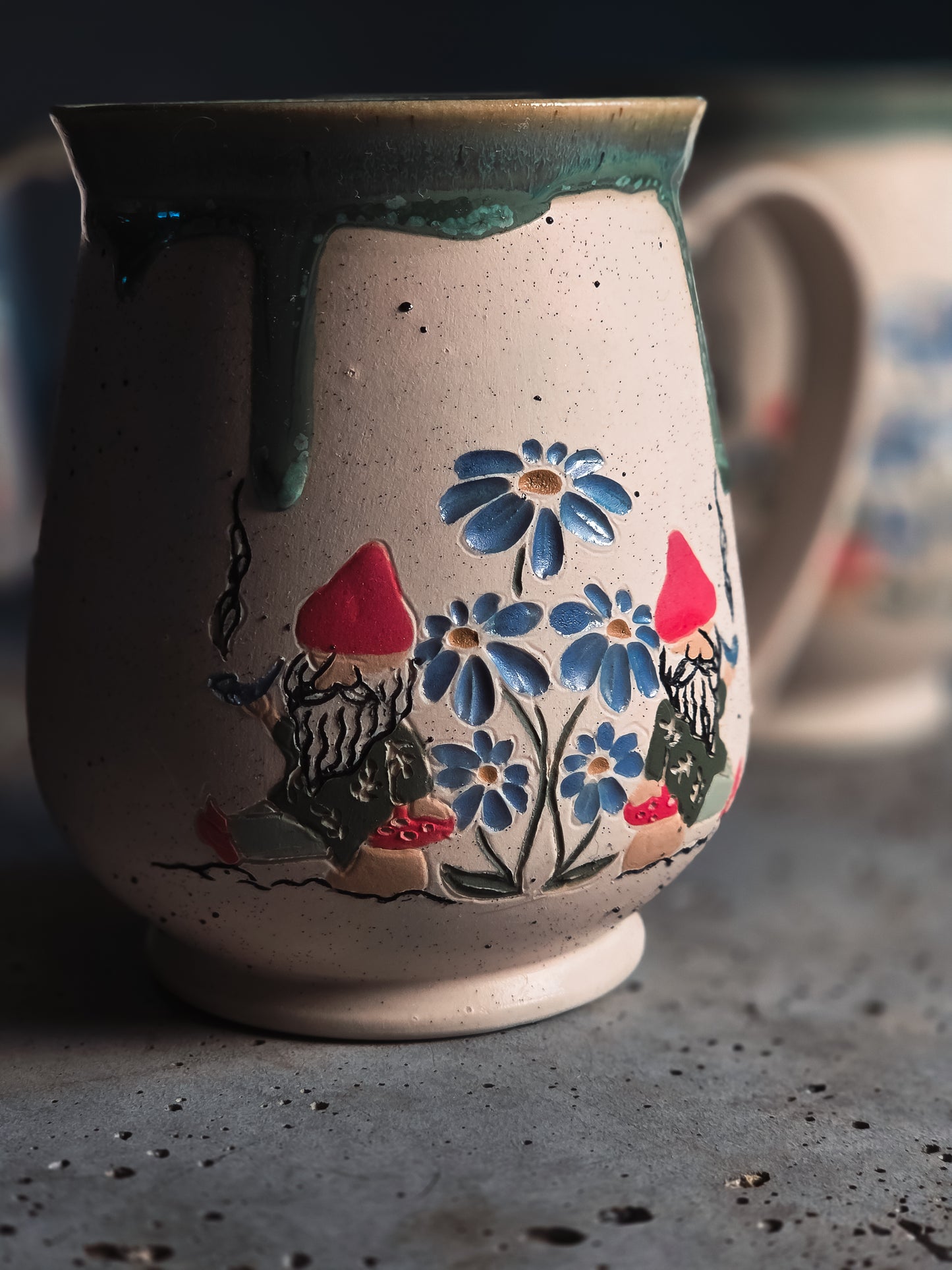 Ceramic Gnome Mug with speckled beige texture, hand-painted blue flowers, and gnomes with red hats under a flowing dark green glaze.
