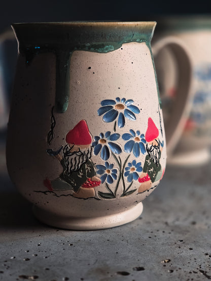 Ceramic Gnome Mug with speckled beige texture, hand-painted blue flowers, and gnomes with red hats under a flowing dark green glaze.