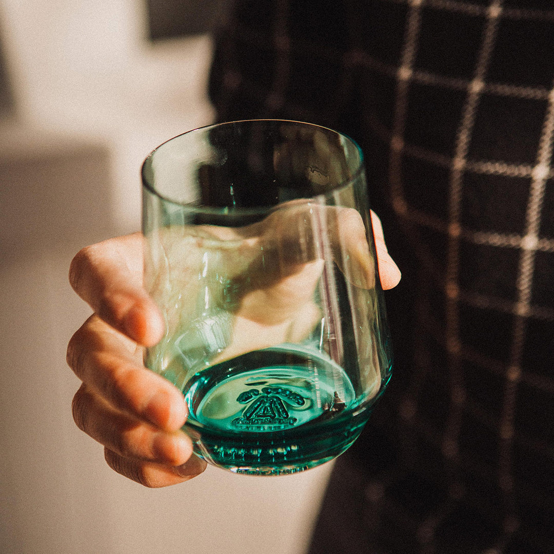 A hand holding a greenish blue tinted wine glass with a Mazama logo stamp at the bottom of the glass.
