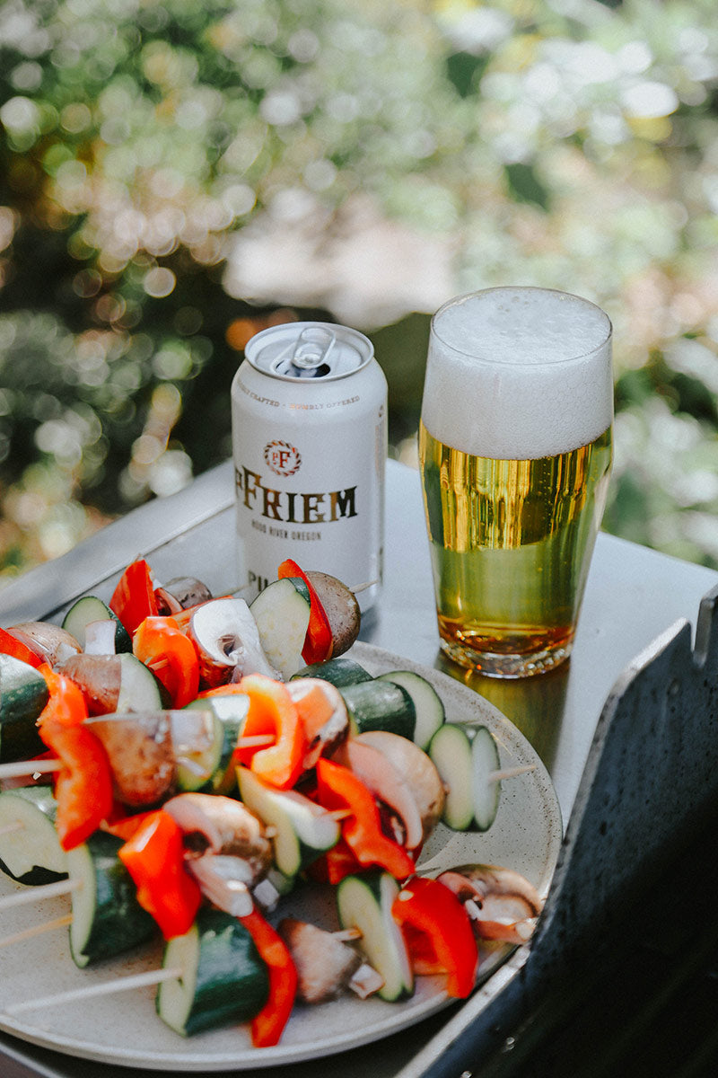 A plate of veggie skewers, can of Pfriem beer and a Mazama pint glass filled with beer on a BBQ grill side table.