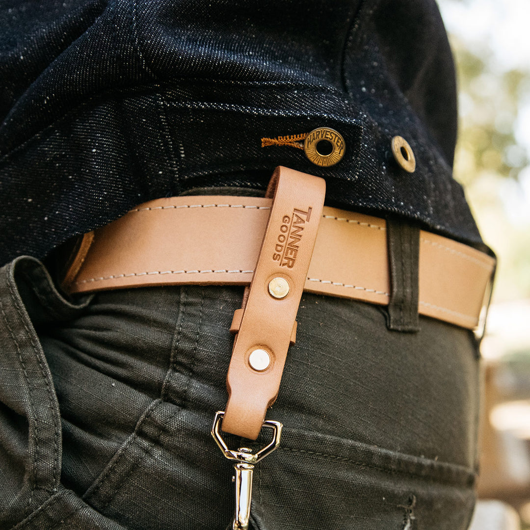 A Tanner Goods leather key lanyard hanging from a natural leather belt on a person's waist. 