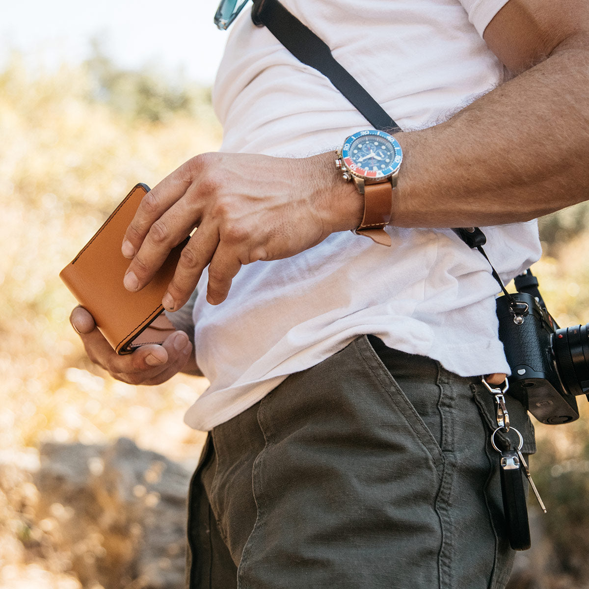 A man opening his saddle tan Bifold wallet with a black leather SLR camera around his chest. 