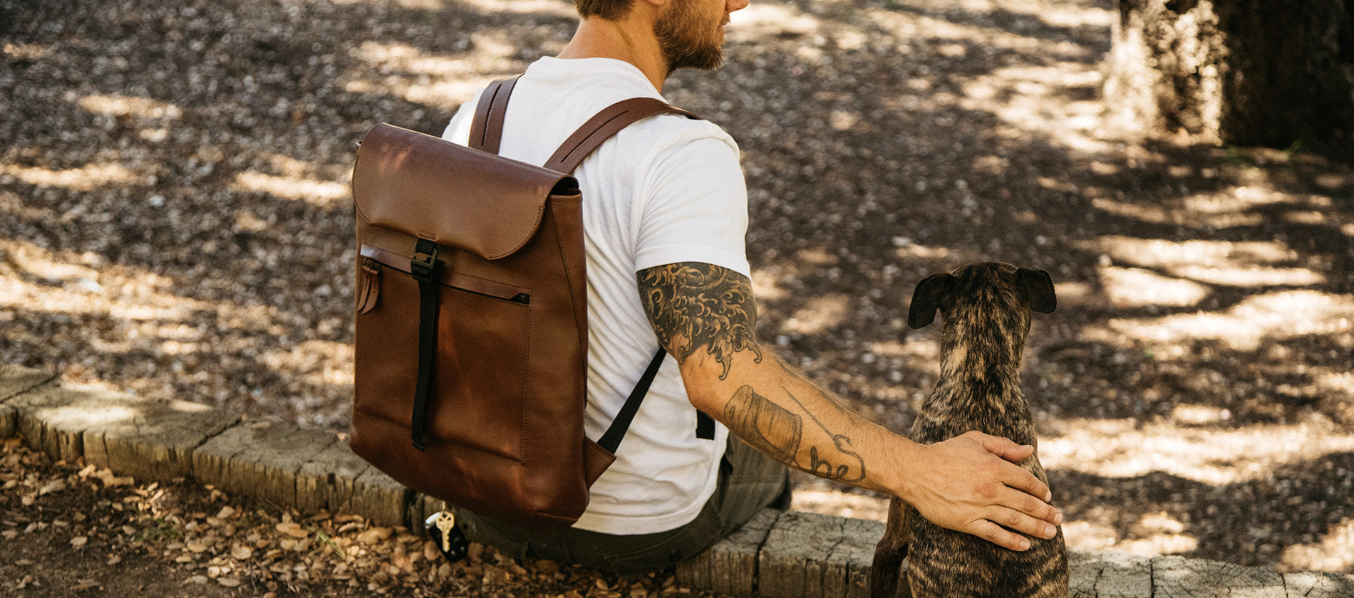 A man sitting down on a hike, petting his brindle patterned dog, wearing a brown all-leather backpack. 