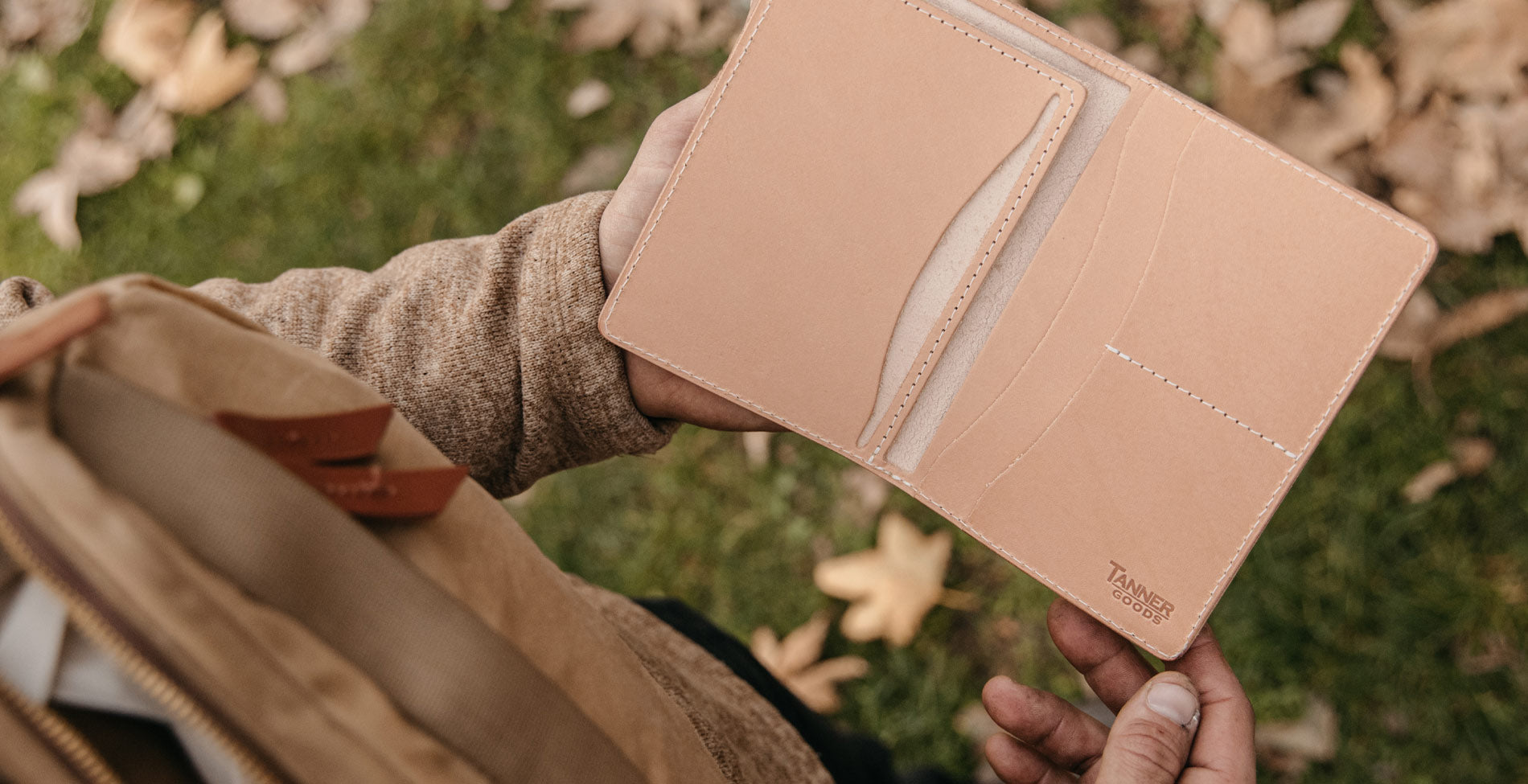 A Travel Wallet in natural leather being opened outside - the color of the leather matching the leaves on the grass.