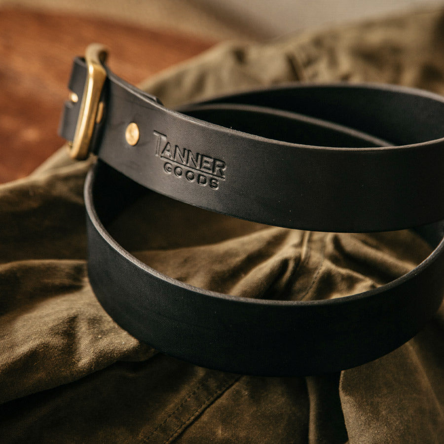 A coiled up black leather belt with Tanner Goods monogram and brass buckle.