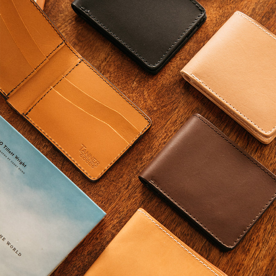Multiple Bifold wallets including a Saddle Tan that is open on top of a coffee table next to a coffee table type book.