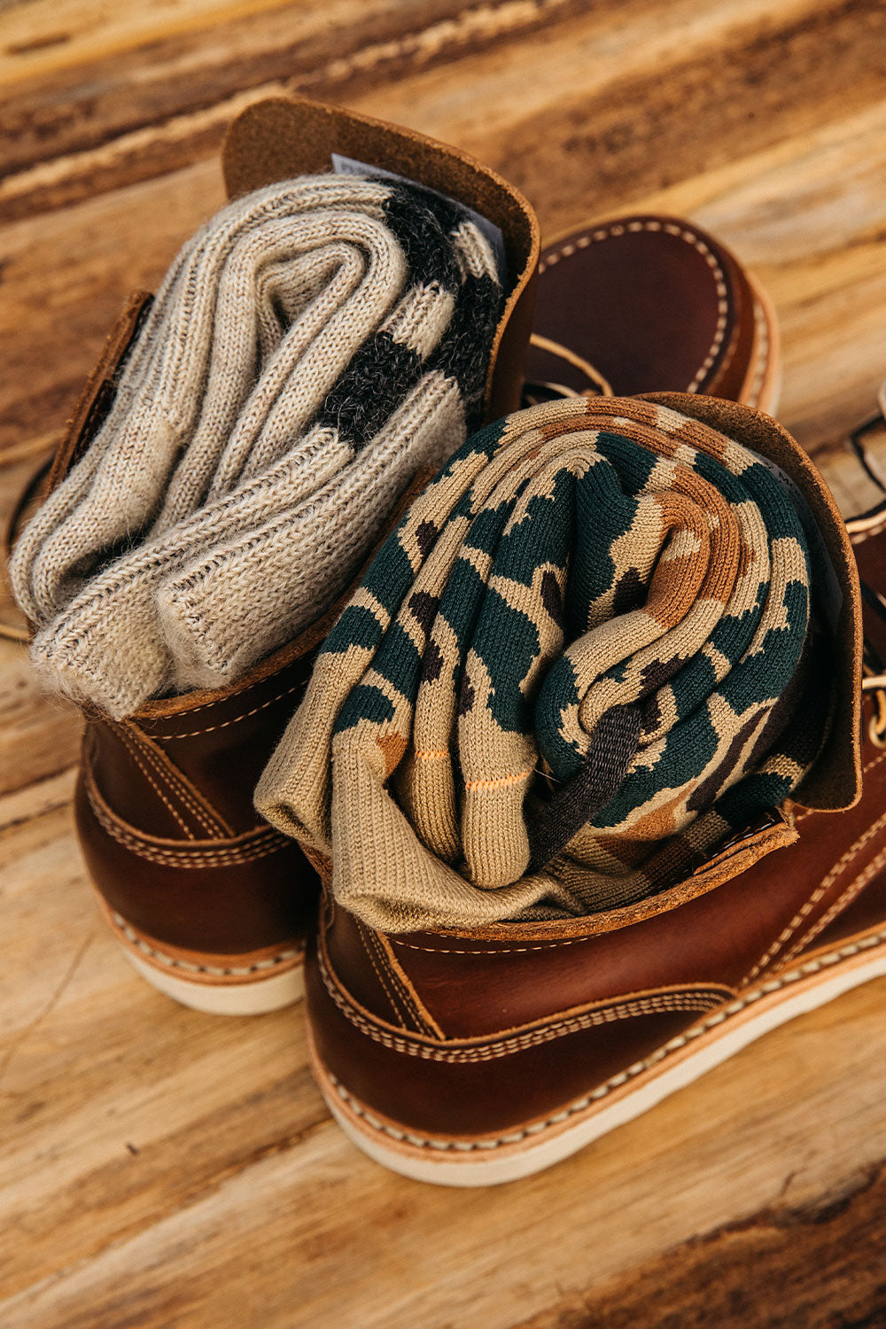 Khaki pile camo crew socks with green and brown patterns, rolled in brown leather boots, next to grey knit socks.