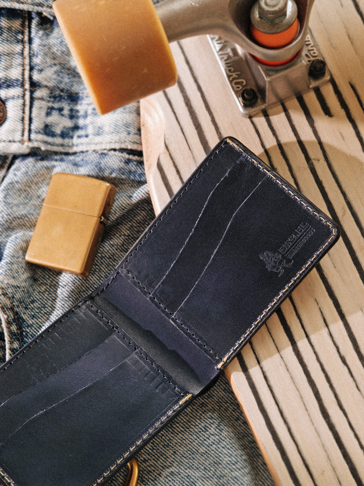 An open blue leather Bifold wallet on top of a pair of jeans, skateboard and golden lighter. 